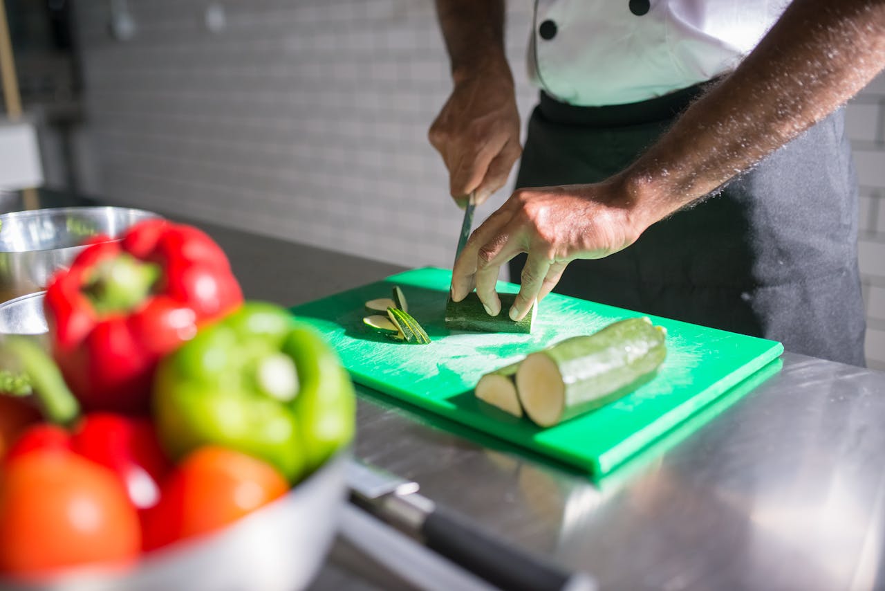our-services-2 Chef slicing zucchini on a green chopping board with fresh vegetables nearby.